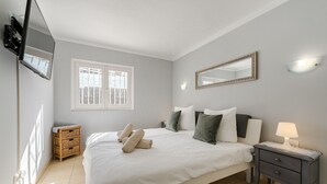 A bedroom with a large bed, a bedside table with a lamp, a small wooden side table, a mirror, and a window with a view of the outside.