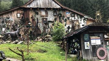 Ein rustikales Holzhaus mit Strohdach, eingebettet in eine natürliche Landschaft mit Bäumen und einem kleinen Schuppen, an dessen Außenwand verschiedene Gegenstände ausgestellt sind.