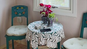 A small room with a round table covered by a white lace tablecloth, two chairs with white cushions, a potted plant with red flowers, and a window with blinds. - 6 Person Holiday Home in Varberg-by Traum (Varberg)