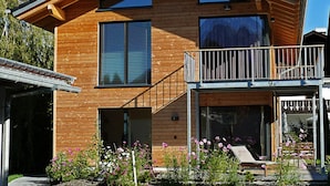 A modern wooden house with a balcony, large windows, and a garden with flowers. - Ferienwohnung Mondholz (Garmisch-Partenkirchen)