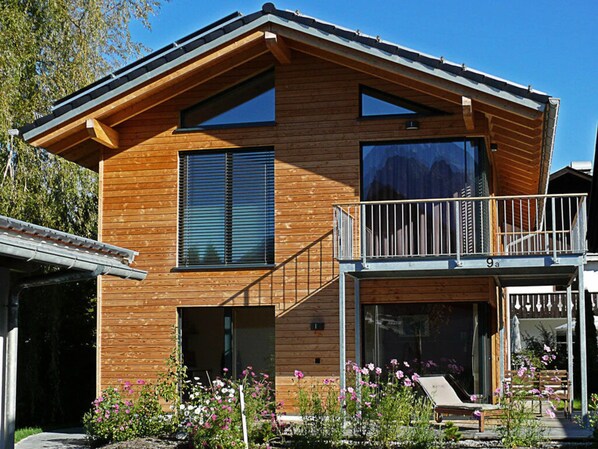 A modern wooden house with a balcony, large windows, and a garden with flowers.