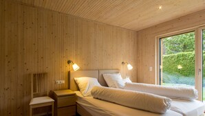 A room with wooden panel walls, a bed with white bedding, a nightstand, a chair, and two bedside lamps. - Ferienwohnung Mondholz (Garmisch-Partenkirchen)