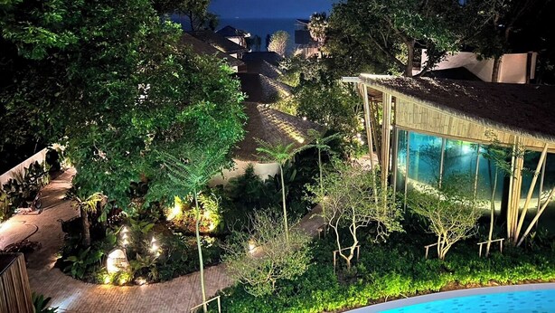 Un espace aménagé au bord de la piscine, avec une piscine entourée de parois en verre, le tout noyé dans une végétation luxuriante et bordé d’arbres.