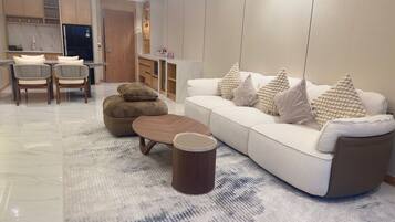 A modern living room with a sofa, ottoman, coffee table, and a small wooden side table. There is a kitchen area in the background with a refrigerator and cabinets.
