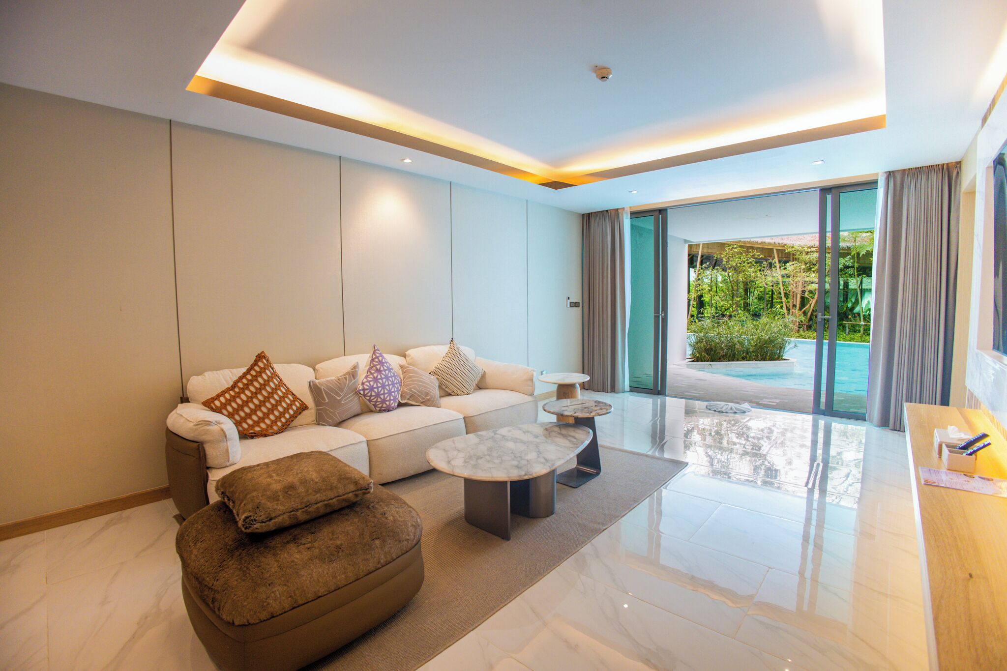 A modern living room with a sofa, ottoman, and coffee table, a glass door leading to a pool area, and a wooden side table.