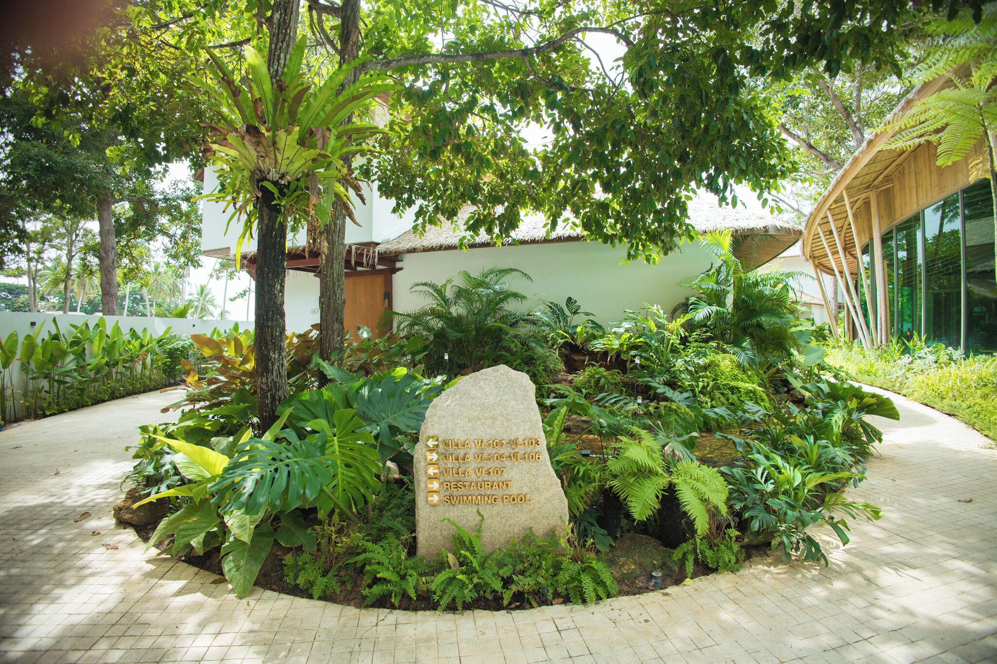 A stone sign with directions to Villa V107, Villa V104, Villa V107, a restaurant, and a swimming pool, surrounded by lush greenery and a paved walkway.