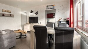 Un salon moderne avec un canapé, une table à manger et des chaises, un téléviseur, et un coin cuisine à l’arrière-plan.