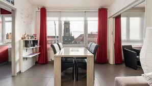 Un salon moderne avec une table à manger, des rideaux rouges et une vue sur le clocher d’une église, visible à travers de grandes fenêtres.