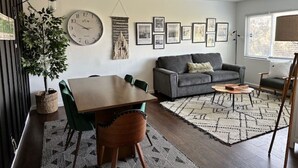 A modern living room with a dining table, sofa, and coffee table, featuring a clock, framed pictures, and a potted plant.