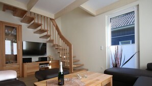 A modern living room with a wooden staircase, a flat-screen TV, a wooden coffee table, and a large window with blinds. - Strandhus Baabe Haus 3 (Ostseebad Baabe)
