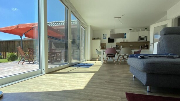 A spacious living area with a large sliding glass door leading to a patio, a grey sofa, and a kitchen area in the background. - Holiday Home on Bornfeld (Osteel)