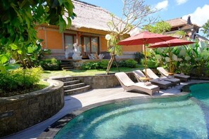 A pool area with lounge chairs and umbrellas, a thatched-roof building, and a well-maintained garden. - PURI ANDONG VILLAS (Ubud)