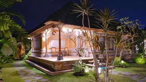 A well-lit wooden building with a patio area, surrounded by lush greenery and stone pathways. - PURI ANDONG VILLAS (Ubud)