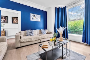 Un salon avec un mur bleu, un canapé beige, une table basse en verre et une fenêtre avec des rideaux.