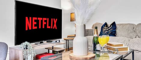 A modern living room with a flat-screen TV displaying 'NETFLIX,' a white vase with white feathers, a bottle of champagne, and two glasses of orange juice on a glass coffee table.