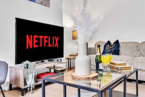 A modern living room with a flat-screen TV displaying 'NETFLIX,' a white vase with white feathers, a bottle of champagne, and two glasses of orange juice on a glass coffee table.