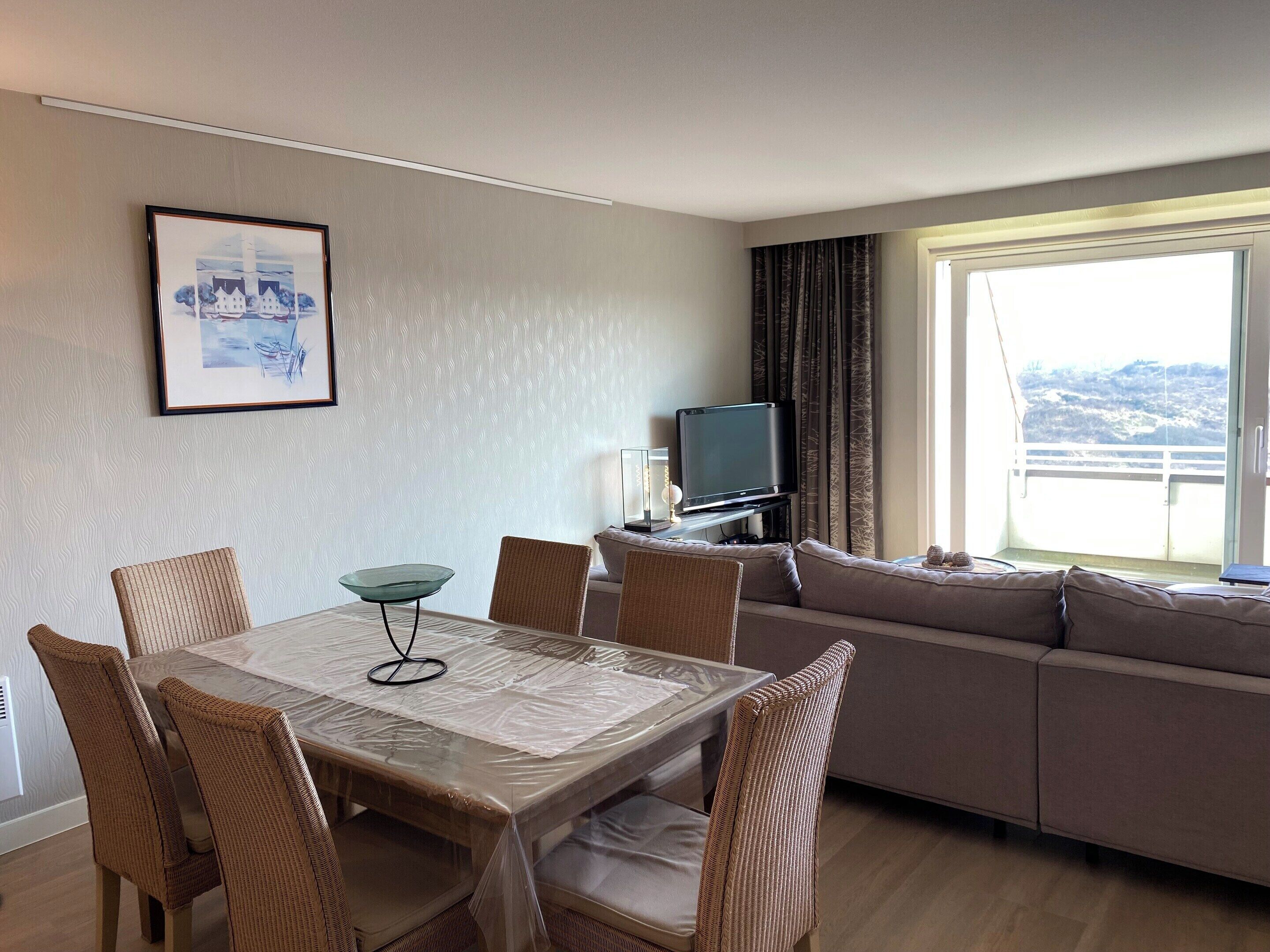 A room with a dining table, chairs, a sofa, a television, and a window with a view.