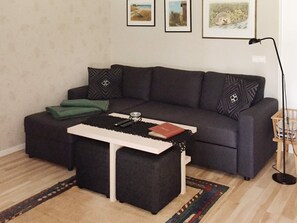 Una sala de estar con un sofá gris oscuro, un reposapiés a juego, una mesa de centro blanca y una alfombra estampada. Hay cuadros enmarcados en la pared y una lámpara de pie.