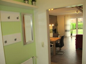 A room with a green wall, a wooden dining table with chairs, a red chair, and a view of a garden through a window. - Holiday Home Heitmann (Dornum)