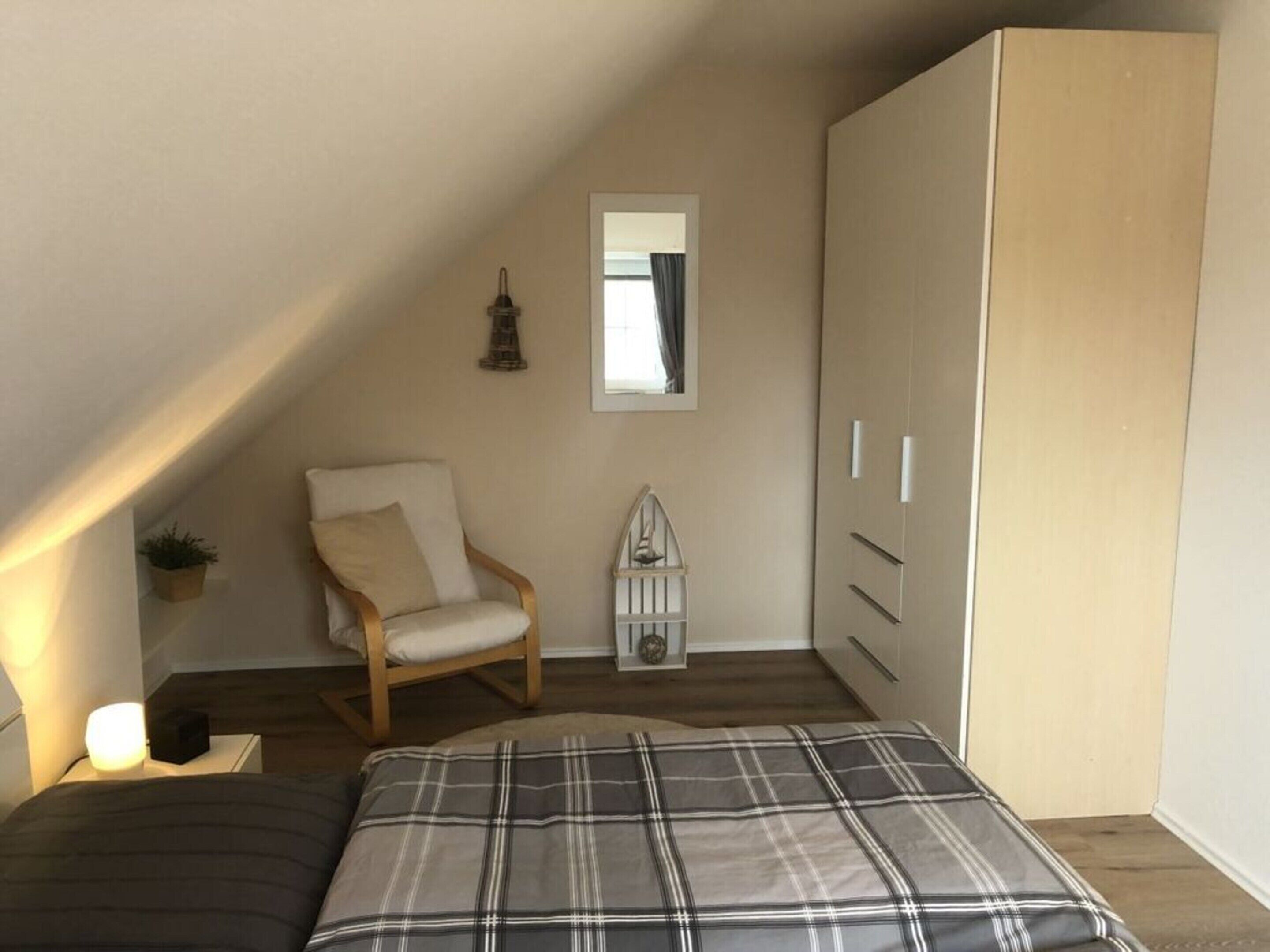 A bedroom with a sloped ceiling, a bed with a plaid bedspread, a wooden chair, a small potted plant, a white cabinet, and a mirror on the wall.