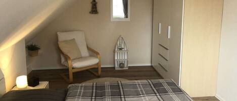 A bedroom with a sloped ceiling, a bed with a plaid bedspread, a wooden chair, a small potted plant, a white cabinet, and a mirror on the wall.