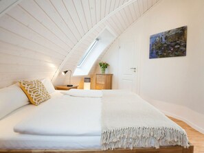A bedroom with a sloped ceiling, a skylight, a bed with white bedding, a bedside table with a lamp, a wooden dresser, and a painting on the wall. - Kugelhaus Phoebe (Penzlin)
