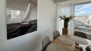A dining area with a large sailboat painting, a potted plant, and a city view through the window. - Cozy Apartment With a top Location (Koksijde)