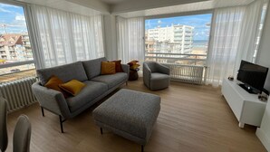 A modern living room with a grey sofa, ottoman, and armchair, a TV on a white cabinet, and large windows with a view of buildings and a beach. - Cozy Apartment With a top Location (Koksijde)