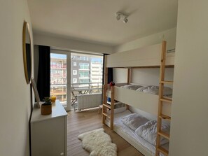 Ein kleines Zimmer mit Etagenbett, Schreibtisch, Stuhl und einem großen Fenster mit Blick auf Gebäude.