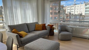 A modern living room with a grey sofa, a matching ottoman, and a grey armchair. The room has large windows offering a view of the cityscape. - Cozy Apartment With a top Location (Koksijde)