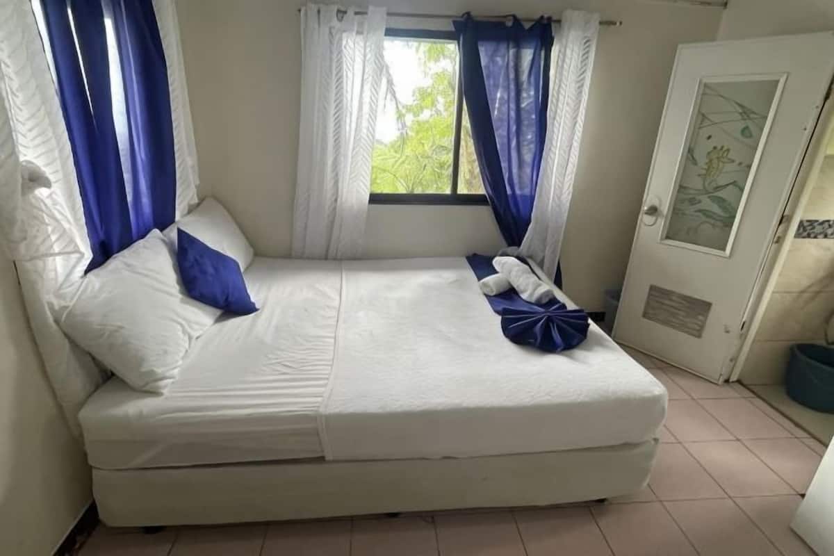 Ein Einzelbett mit weißer Bettwäsche, blauen Kissen und Vorhängen, eine Tür mit Fenster sowie ein gefliester Boden.