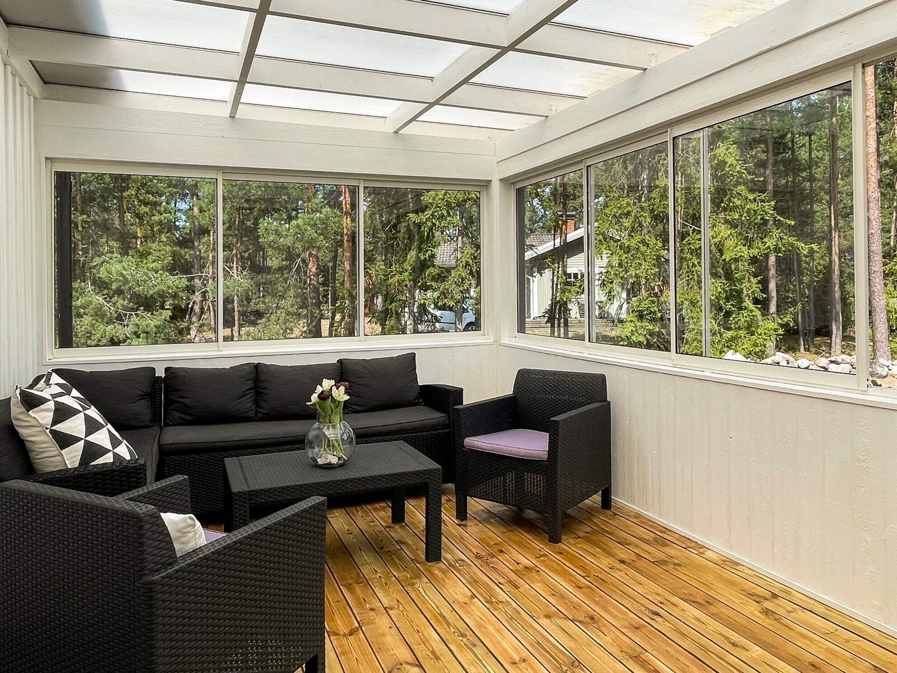 A sunroom with a wooden floor, black wicker furniture, and large windows offering a view of trees.