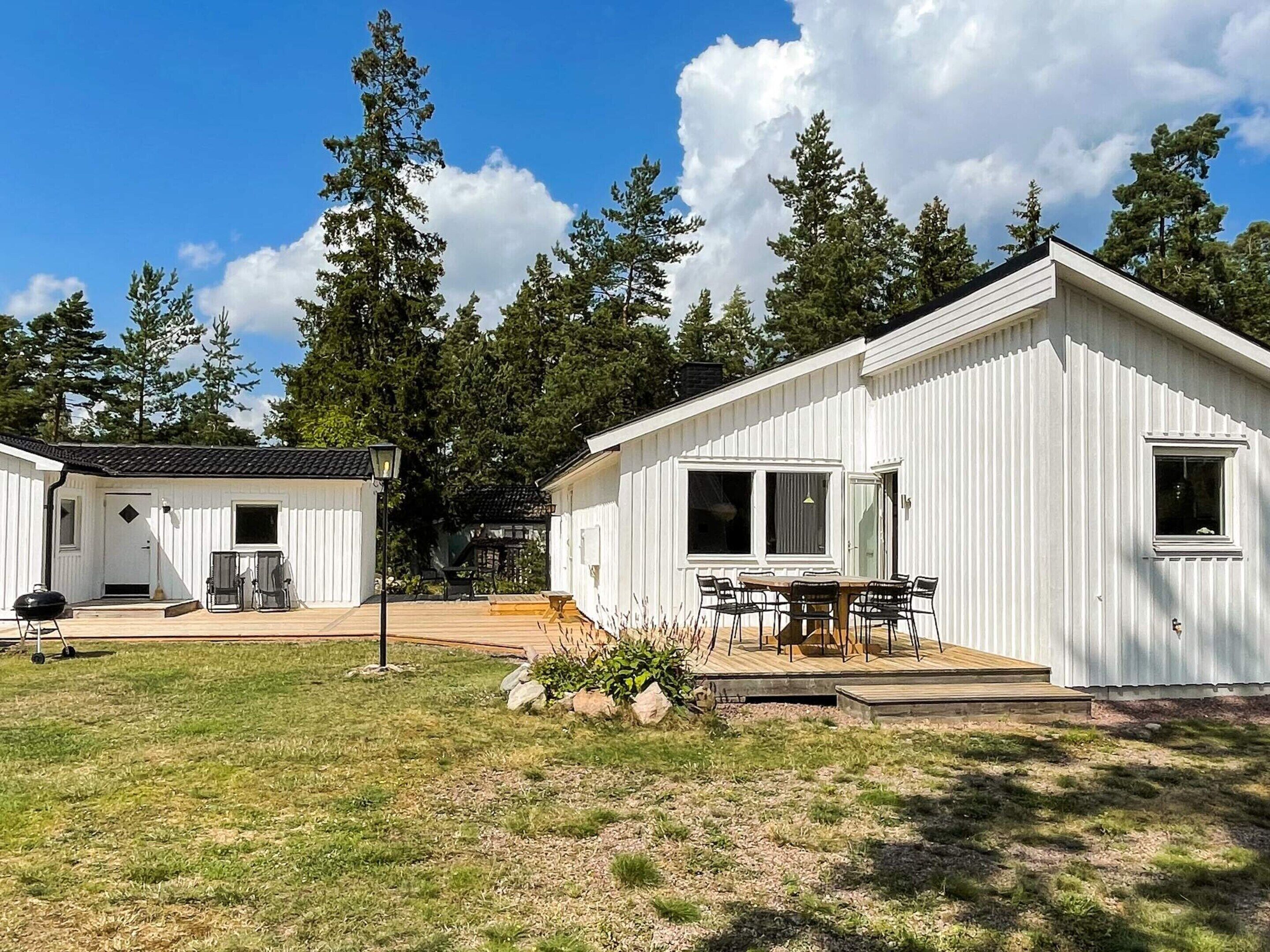 A white cabin with a deck and a dining area, surrounded by trees.