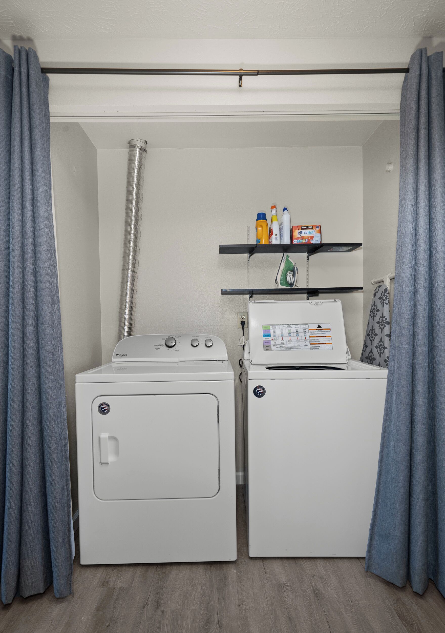 A laundry room with two white washing machines, a vent pipe, and shelves with laundry supplies.