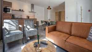 A modern living room with a brown sofa, two grey armchairs, and a wooden coffee table. A kitchen with a microwave and a dining area with a table and chairs is visible in the background. - Luxury new Apartment on Ameland (Buren)