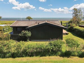 A single-story wooden house with a sloped roof, surrounded by a grassy yard and dense shrubbery.