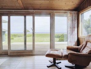 Una habitación con puerta de vidrio corredera que da a una zona verde, un sillón de cuero marrón y una ventana con cortinas.