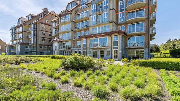 Un edificio residencial de varios pisos con balcones, ventanas amplias y un jardín ajardinado en la parte delantera.