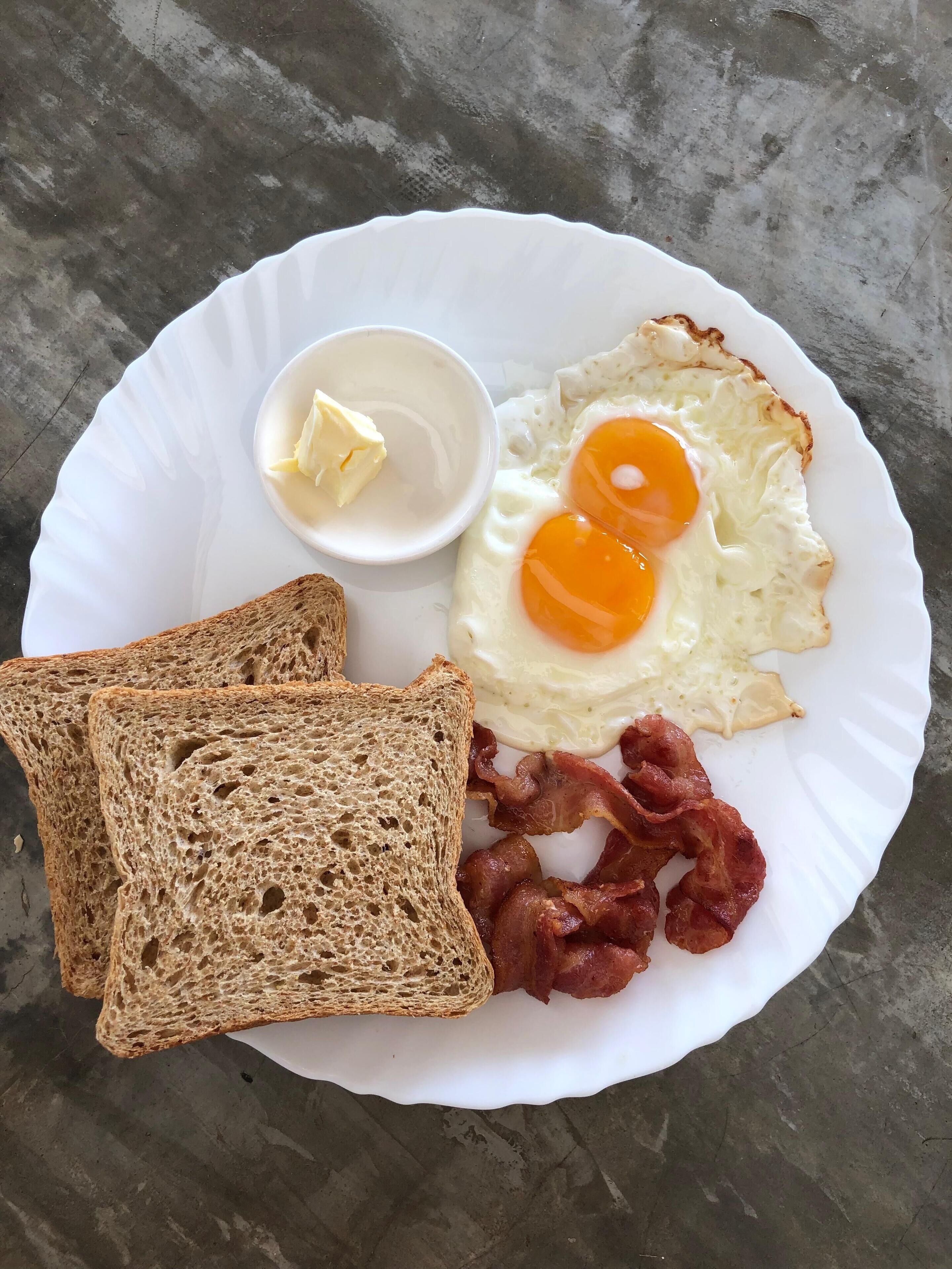 En tallerken med to spejlæg, en stykke bacon, en skive ristet brød og en lille skål smør.
