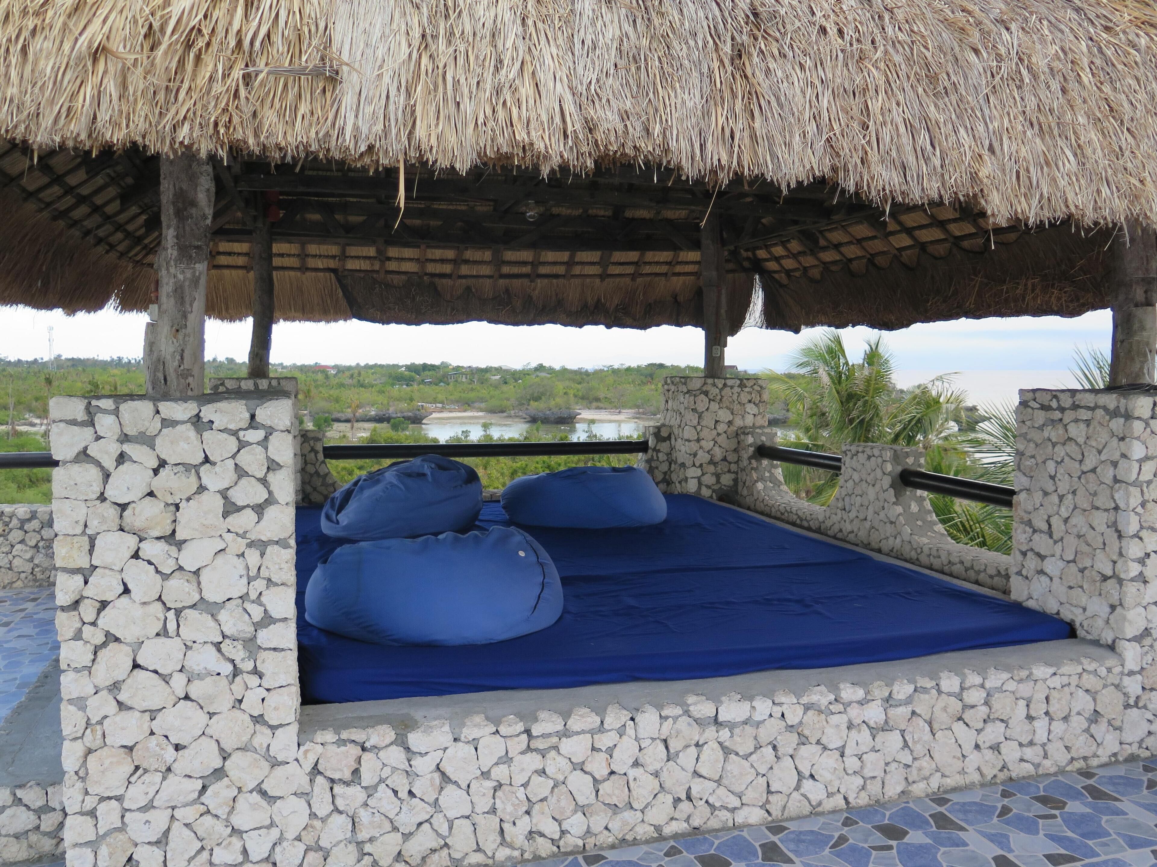 A stone-walled bed with blue bedding, a thatched roof, and a view of a lake and greenery.
