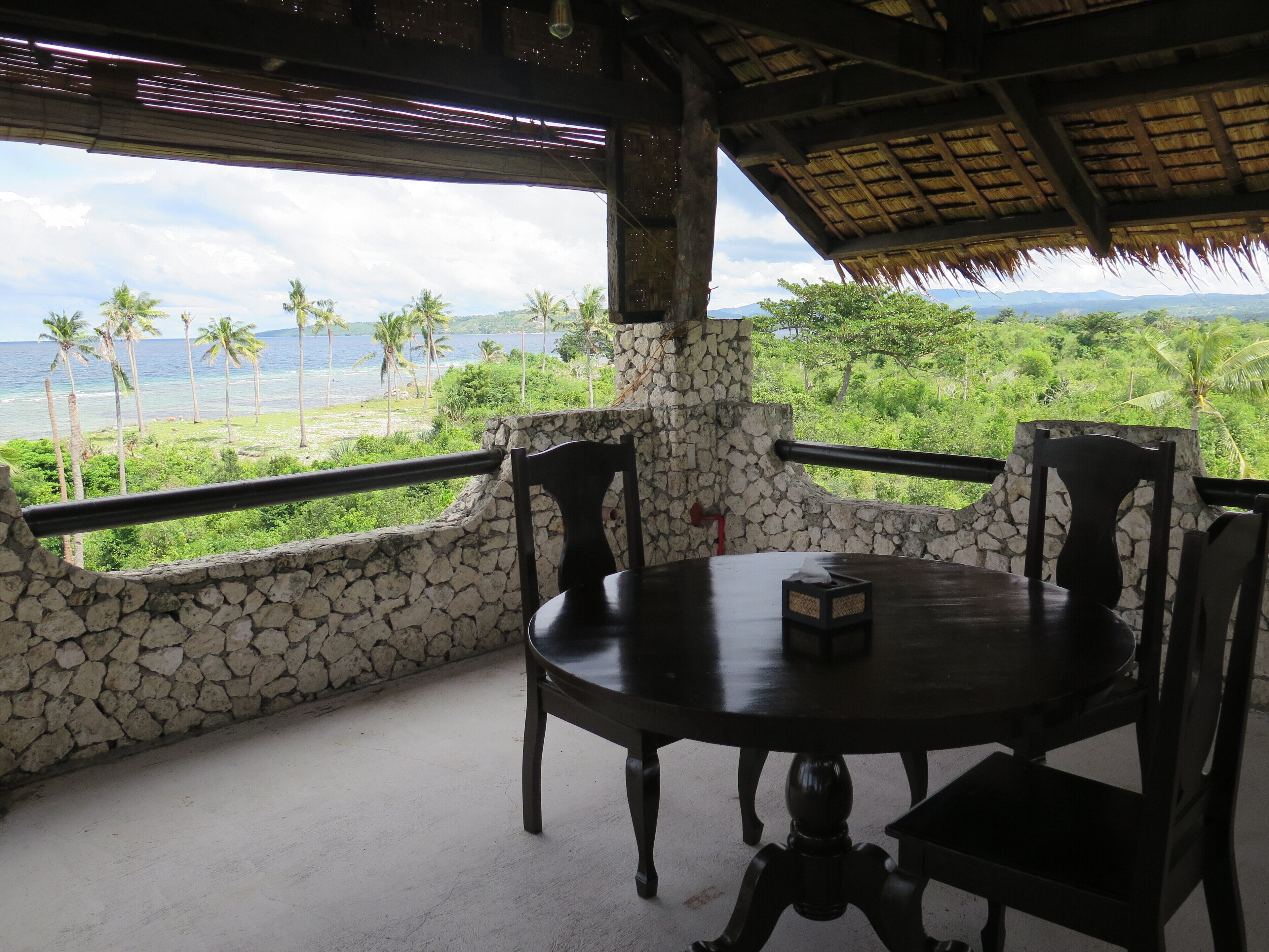 Quarto com paredes de pedra, uma mesa redonda e cadeiras, com vista para uma paisagem exuberante com palmeiras e o oceano.