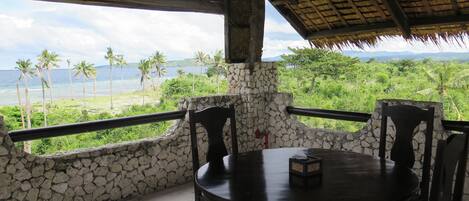 Quarto com paredes de pedra, uma mesa redonda e cadeiras, com vista para uma paisagem exuberante com palmeiras e o oceano.
