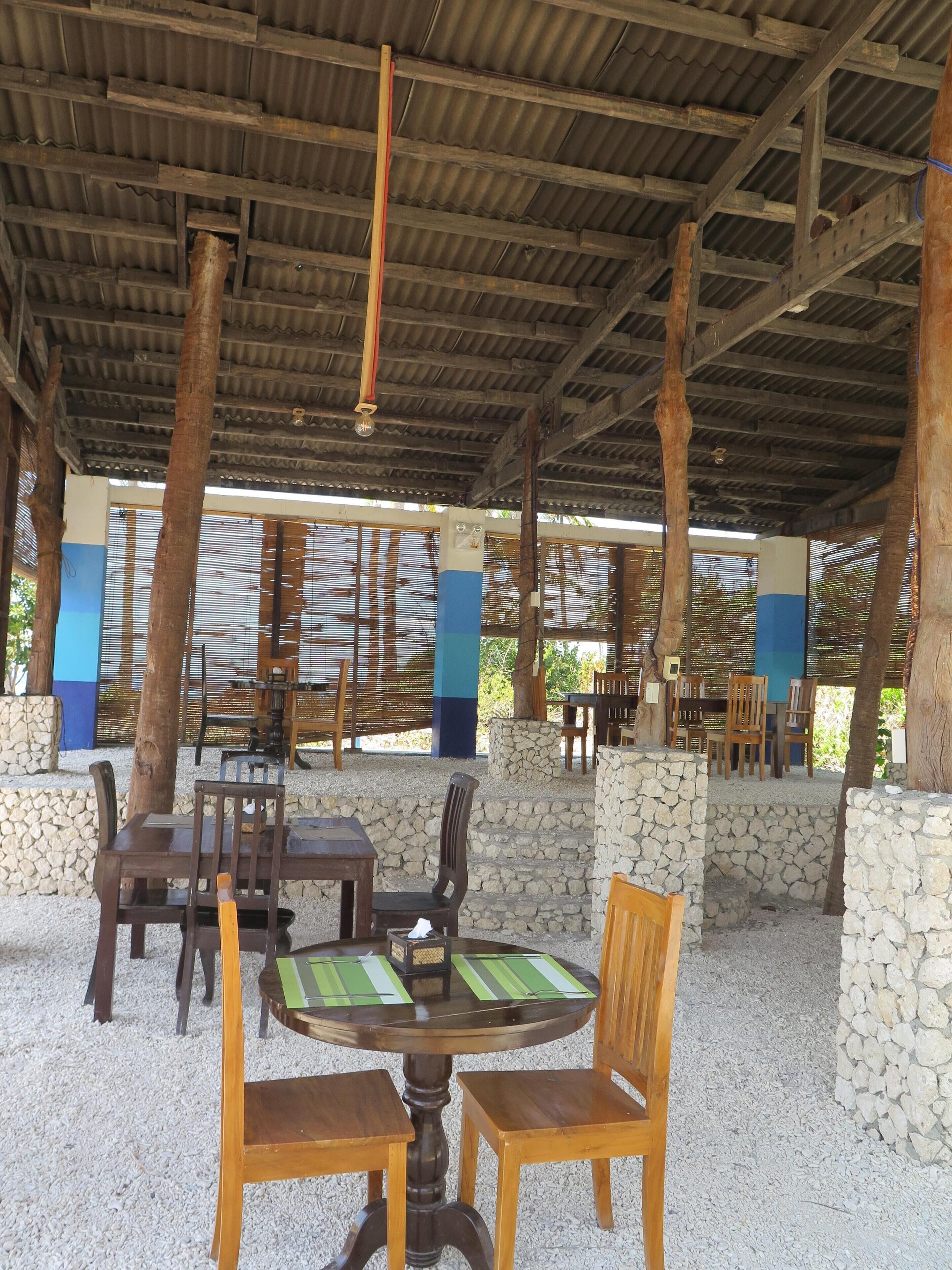 Un espace extérieur couvert, aménagé en patio, avec des tables et des chaises en bois, des murs en pierre et un toit en métal ondulé.
