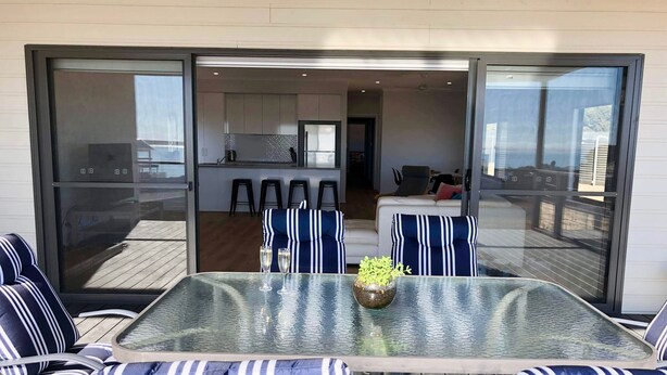 A glass-enclosed outdoor dining area with striped cushions and a glass table, leading to a modern kitchen and a seating area with a sofa and chairs.