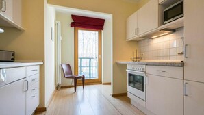 Una cocina moderna con armarios blancos, horno empotrado y microondas. Hay una silla cerca de una ventana con un cortinero rojo.
