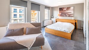 Quarto moderno com sofá, cama de madeira, janela ampla e uma obra de arte com tema praiano na parede.