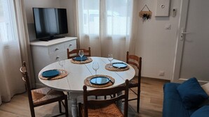 Un'area pranzo con un tavolo rotondo apparecchiato per quattro persone, circondato da sedie di legno con sedili intrecciati. Un televisore è montato sulla parete sopra il tavolo.