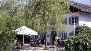 An outdoor seating area with tables and chairs under a white umbrella, surrounded by trees and a building with blue shutters.