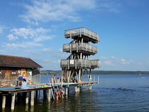 A wooden pier with a multi-level wooden structure extending into the water, a building with a sign, and people swimming and relaxing. - Schleichhof Ammersee (Dießen am Ammersee)