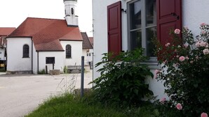 A white building with red shutters and a window, surrounded by greenery and a church with a red roof in the background.
